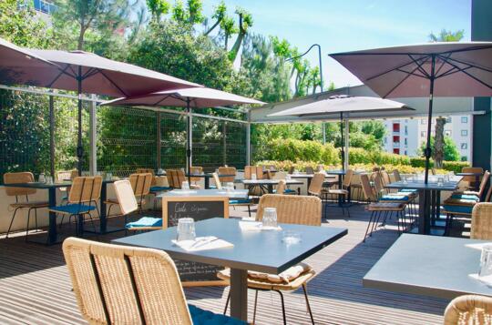 Terraza soleada en Bistrot City de Appart'City, con mobiliario cómodo bajo sombrillas, escenario perfecto para un almuerzo al aire libre o relajación.