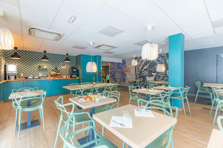 Dining room of a Clef Verte certified aparthotel with light wood chairs and tables, modern white pendant lights, a tiled bar counter, blue-grey walls, and a foliage mural, creating a contemporary and eco-friendly ambiance.