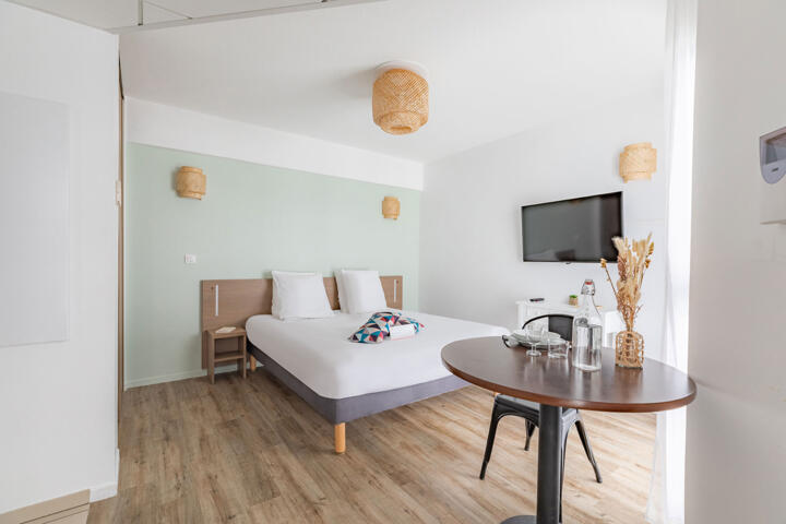 Helles und minimalistisches Aparthotel-Zimmer mit Clef Verte Zertifizierung, mit einem Doppelbett, hängenden Korblampen, einem Flachbildfernseher an der Wand, einem kleinen Essbereich mit einem runden Tisch und natürlichen Dekorationselementen, was einen umweltfreundlichen Ansatz in der Unterkunft widerspiegelt.