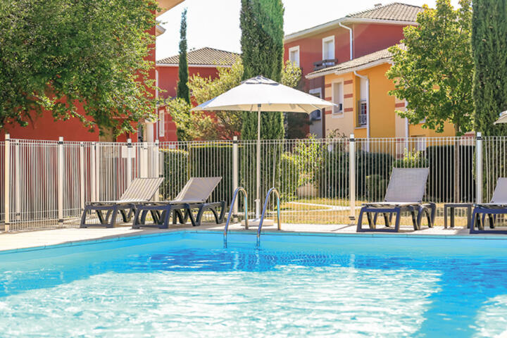 Piscine extérieure ensoleillée chez Appart'City, entourée d'une clôture de sécurité, avec des transats confortables sous un parasol blanc, offrant un espace de détente pour les familles en séjour.