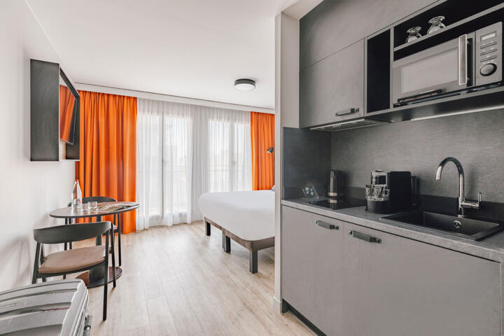 A bright and welcoming Appart'City aparthotel room with a comfortable double bed, bright orange curtains, a wooden dining table, matching chairs, and a modern, well-equipped kitchenette, offering an ideal and convenient place to stay for visitors to the sporting events in France in 2024.