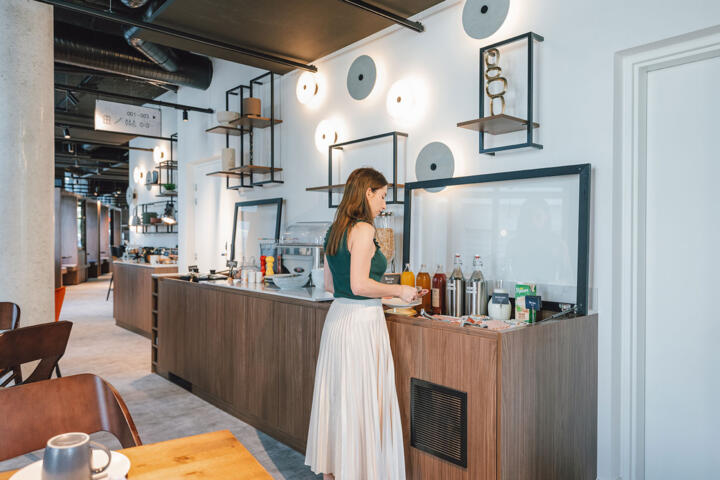 An elegant woman serves herself breakfast in the modern, well-lit dining area of a Parisian apartment hotel, with its designer interior and welcoming atmosphere.