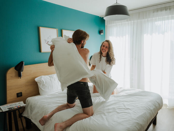Una pareja sonriente mantiene una pelea de almohadas en una luminosa habitación de apartahotel con decoración moderna y ambiente relajado, símbolo de una estancia feliz y despreocupada.