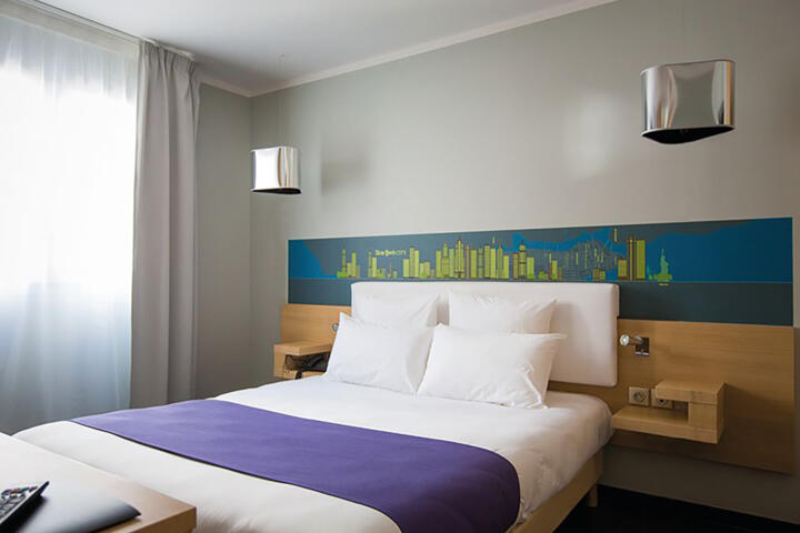 Zeitgenössisches Appart'City Aparthotel-Zimmer mit einem großen Doppelbett, pflaumenfarbener Bettwäsche, flauschigen Kissen und einem künstlerischen Kopfteil mit einer städtischen Skyline, für einen städtischen Aufenthalt mit Komfort und Stil.