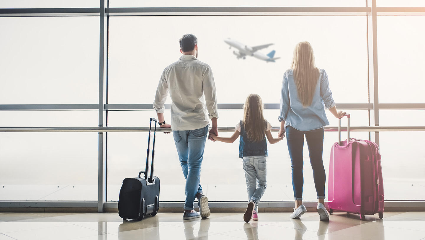 Family watching a plane take off at Strasbourg Airport, holding hands with luggage