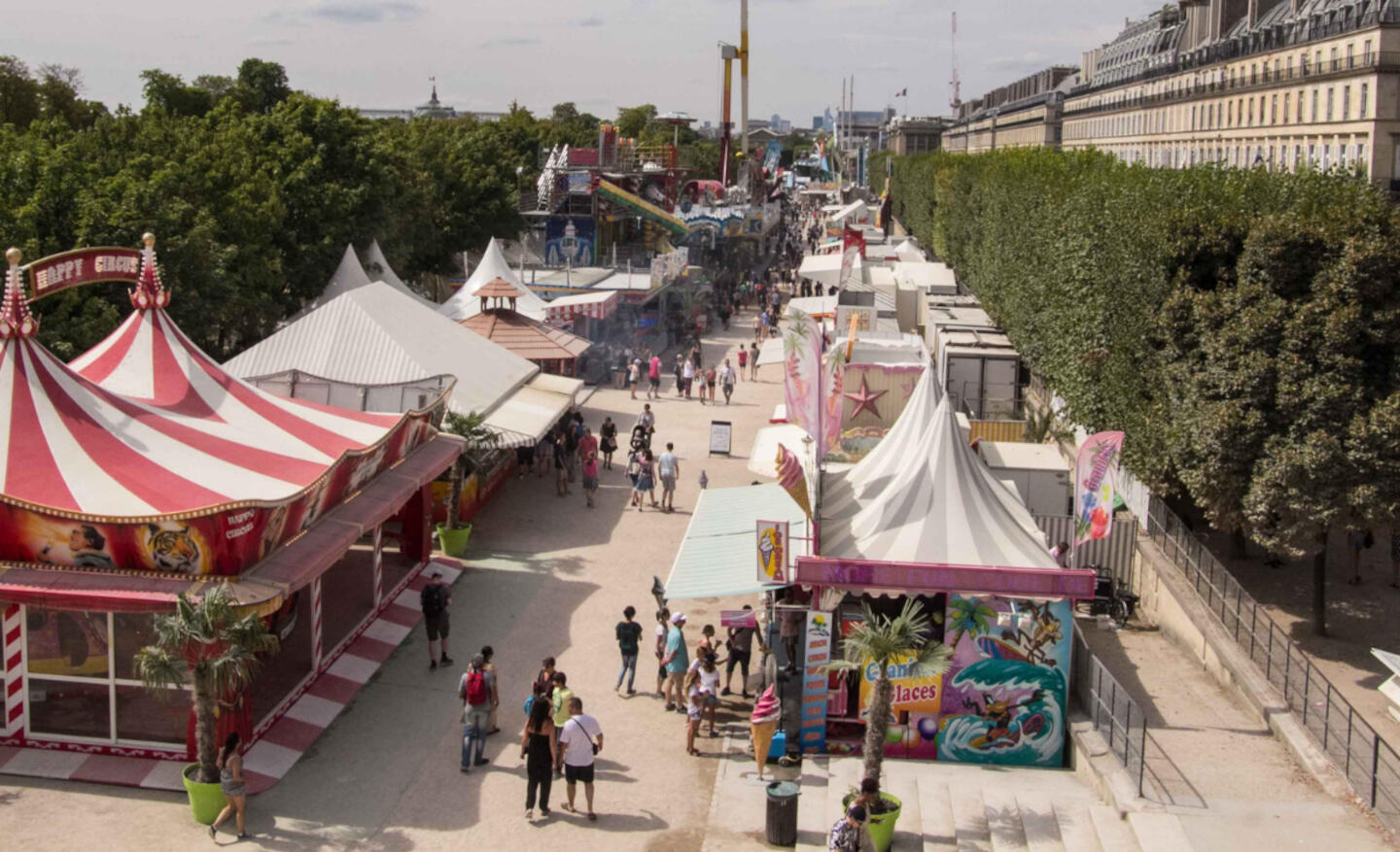 Allée centrale de la Fête des Tuileries avec stands colorés et visiteurs profitant des attractions sous un ciel bleu clair.