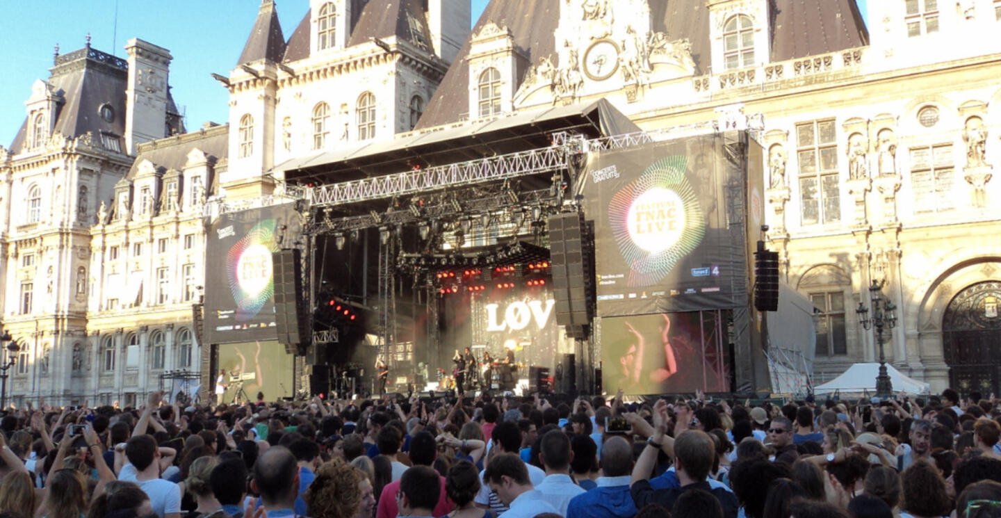 Crowd enjoying a live concert at the FNAC Music Festival in Paris, in front of a historic building with the stage presenting the performance 'LOVE' under a clear blue sky.