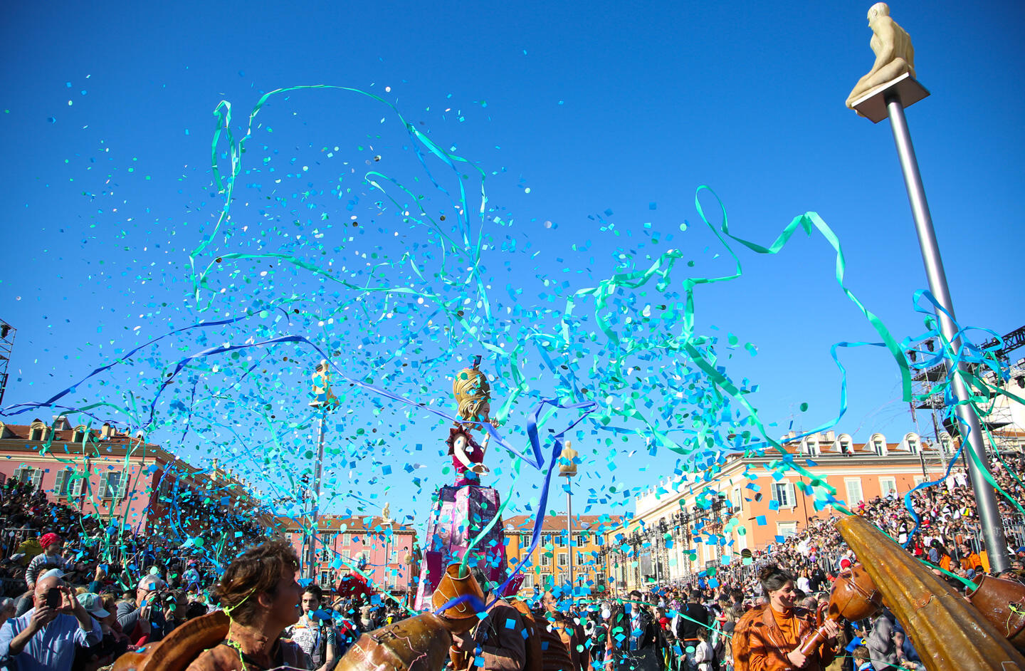 Celebración animada del Carnaval de Niza con confeti turquesa volando en el aire, multitud entusiasta y coloridas esculturas de carnaval bajo el cielo azul.