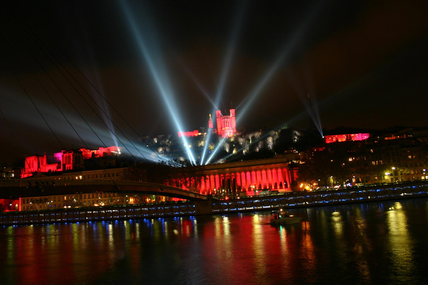 Night view of Lyon, with red illuminations of the Basilique de Fourvière and the historic Palais de Justice, beams of light crossing the sky above the bridge, and reflections of the lights on the Saône during the Fête des Lumières.