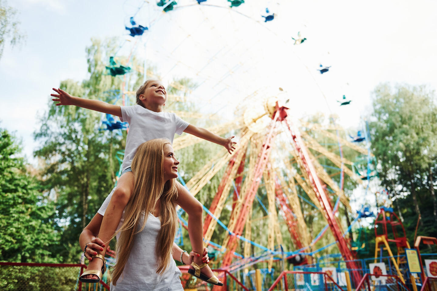 Lächelndes Kind auf den Schultern einer Frau, Arme in die Luft streckend mit einem farbenfrohen Riesenrad im Hintergrund, das Glück und Spaß im Antibes Land Park symbolisiert.