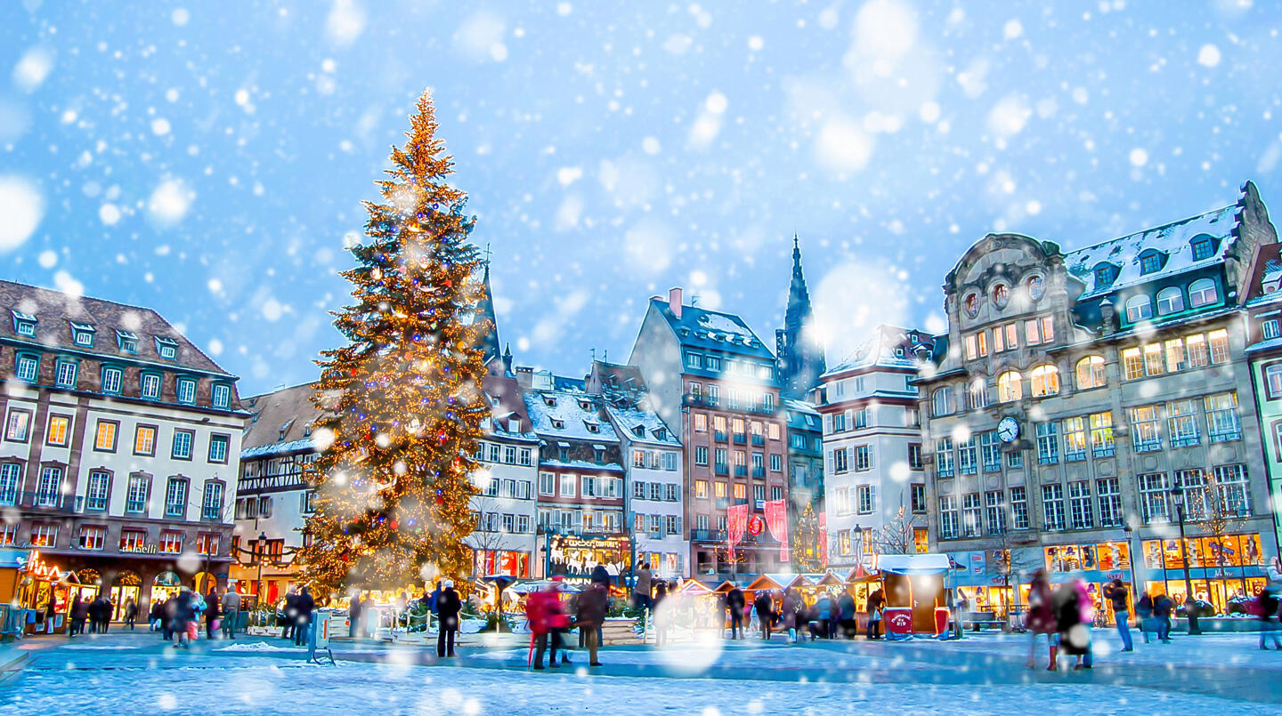A lively Christmas market with a large illuminated fir tree and snow-covered visitors, ideal for festive city breaks.