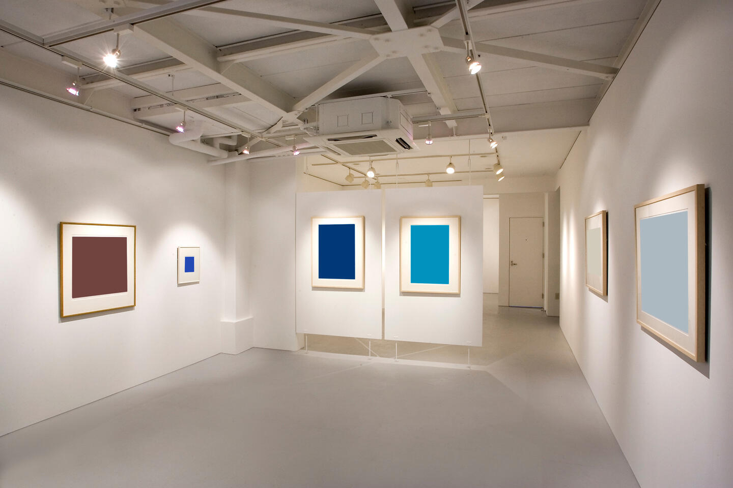 Interior minimalista de una galería de arte moderno en la Feria Internacional de Fotografía Artística de París, mostrando una serie de fotografías enmarcadas de colores vivos en paredes blancas iluminadas por focos. El entorno limpio resalta las obras expuestas.