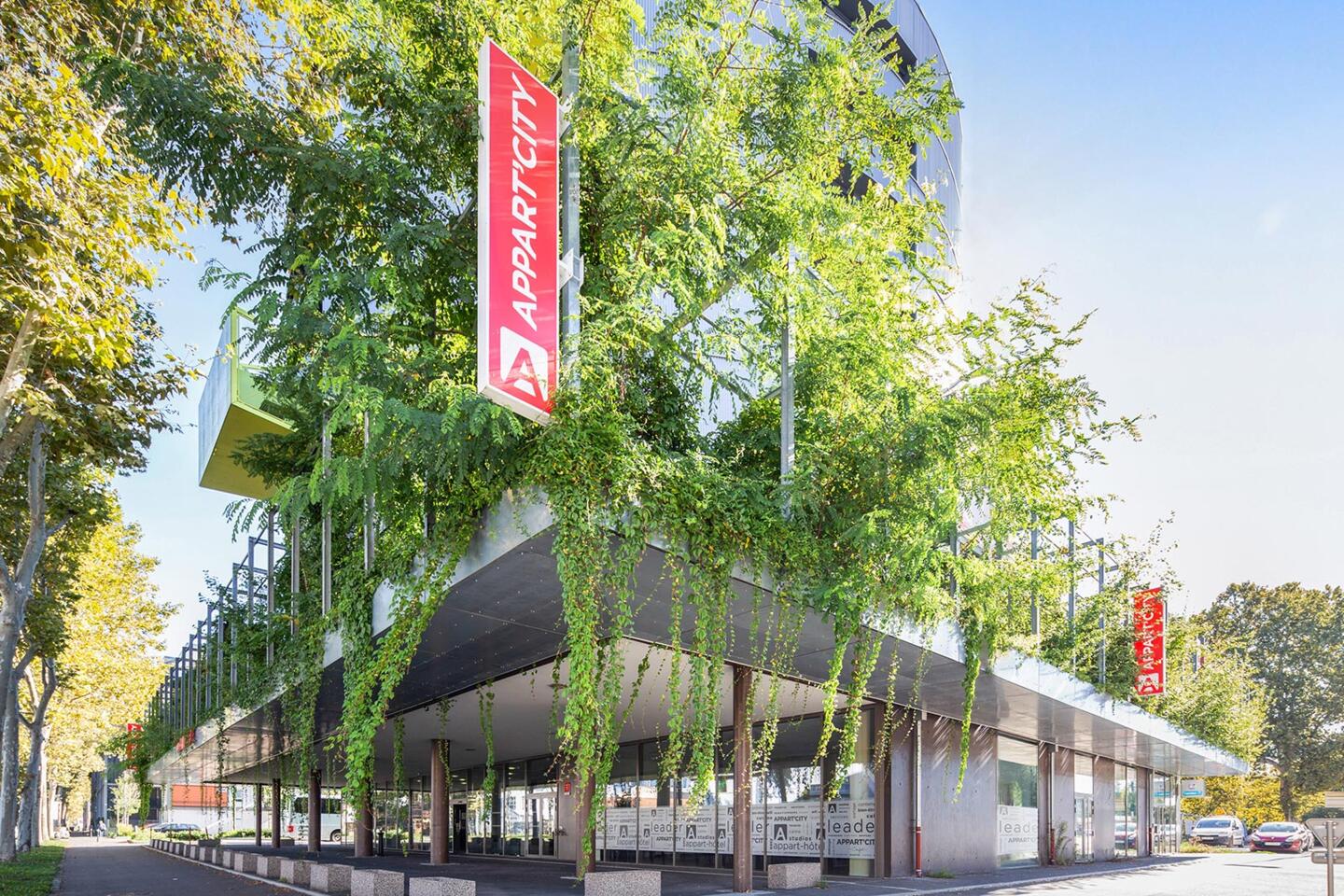 Außenfassade des Appart'City mit vertikalen roten Schildern, gelegen an einer baumbestandenen Straße, zeigt ein modernes umweltfreundliches Aparthotel in städtischer Umgebung.