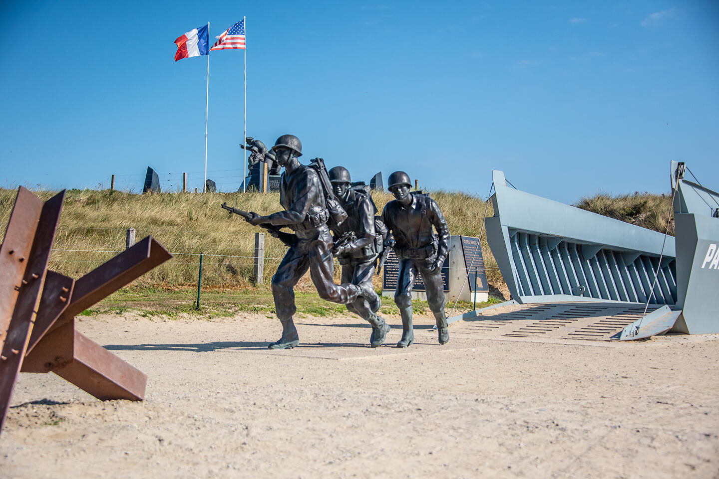 Statuen von vorrückenden Soldaten, die die D-Day-Landungen am Omaha Beach in der Normandie, Frankreich, darstellen. Hinter den Statuen wehen die französischen und amerikanischen Flaggen, und eine Nachbildung eines Landungsbootes, das während der Invasion der Normandie verwendet wurde.