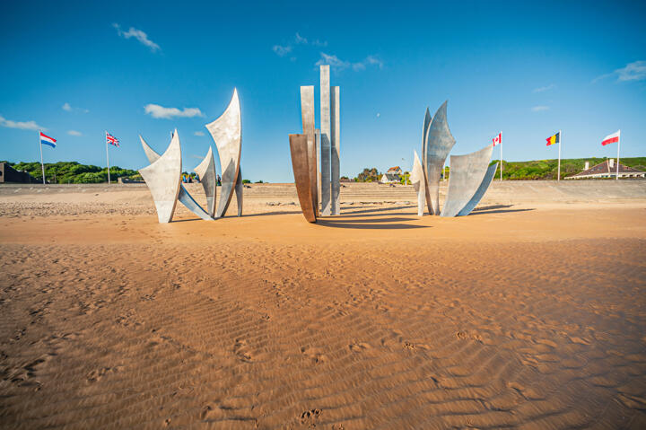 Les Braves-Denkmal am Omaha Beach in der Normandie, Frankreich. Diese moderne Skulptur, die die Soldaten der D-Day-Landungen ehrt, steht im Sand mit den Flaggen mehrerer alliierter Länder im Hintergrund unter einem klaren blauen Himmel.