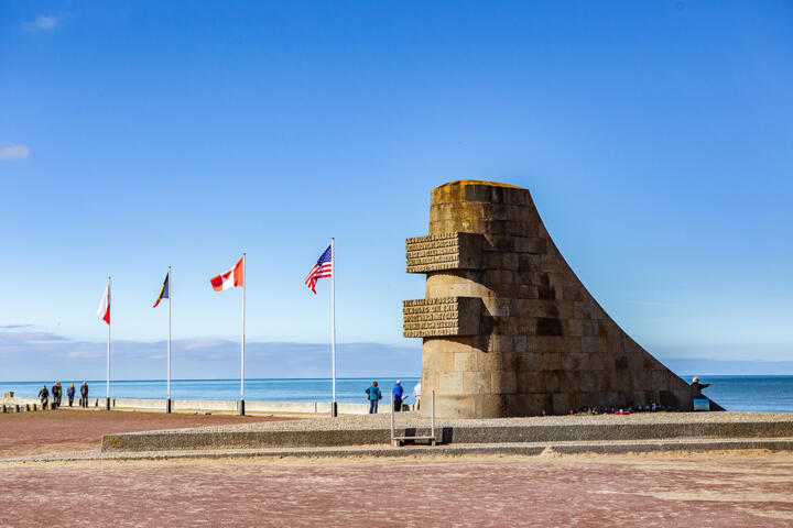 Gedenkdenkmal am Juno Beach in der Normandie, Frankreich, mit Flaggen mehrerer alliierter Länder, darunter Polen, Kanada und die Vereinigten Staaten, die im Hintergrund unter einem klaren blauen Himmel wehen.