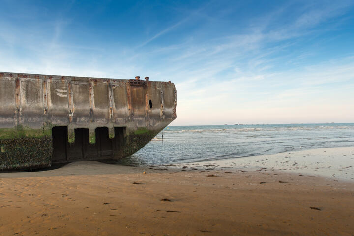 Blick auf ein Überbleibsel eines künstlichen Hafens am Strand von Arromanches in der Normandie, Frankreich. Dieser Hafen, der während der D-Day-Landungen genutzt wurde, liegt bei Ebbe auf dem Sand, mit dem Meer im Hintergrund unter einem blauen Himmel.