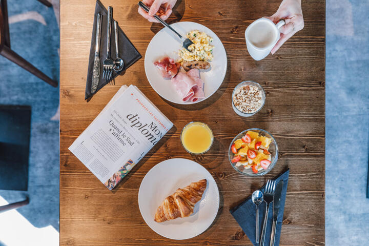 Draufsicht auf ein Frühstück mit einem Croissant, Rührei, Schinken, frischem Obst, Müsli und einem Glas Orangensaft, mit einer Zeitung auf dem Tisch, im Aparthotel Paris Grande Bibliothèque.