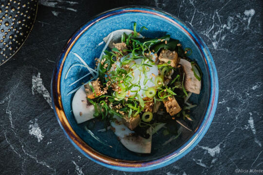 Plat servi dans un bol bleu, composé de tofu mariné, d'œuf mollet, de légumes frais, d'herbes et de graines de sésame, présenté sur une table en marbre noir.