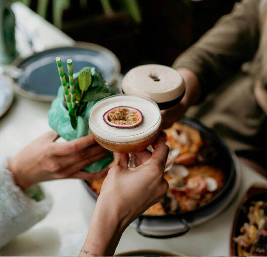 Gros plan sur deux cocktails festifs : un cocktail exotique avec glaçons, menthe et pailles vertes, et un autre avec une mousse garnie d’un demi-fruit de la passion, tenus par deux personnes en train de trinquer.