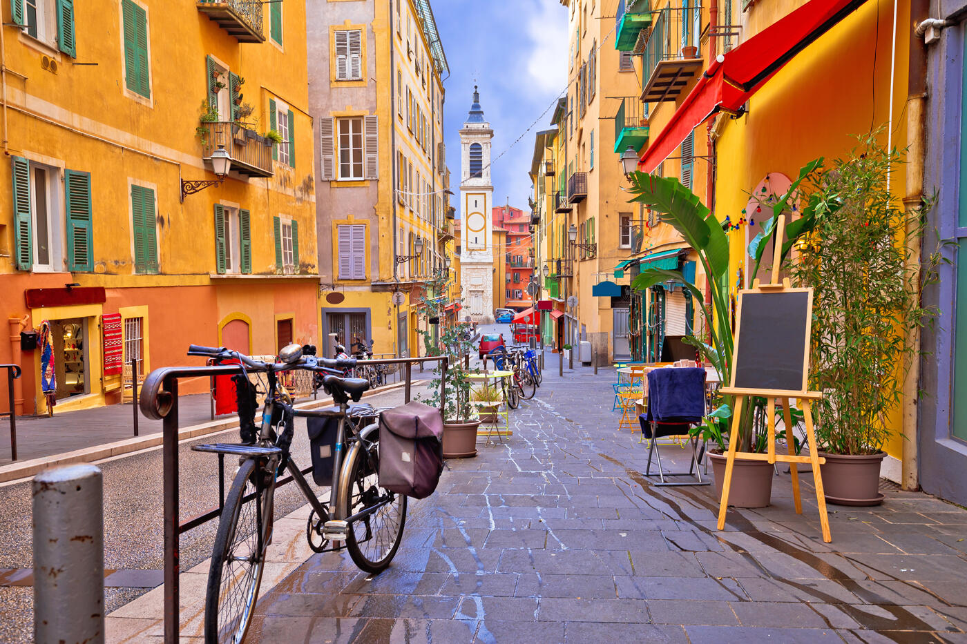 Malerische Straße in der Altstadt von Nizza mit bunten Gebäuden, Straßencafés, einem geparkten Fahrrad und einem Glockenturm im Hintergrund