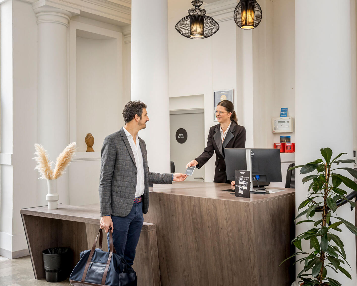 Homme arrivant à la réception d’un établissement, recevant une carte de chambre d’une réceptionniste souriante derrière un comptoir moderne, dans un intérieur lumineux aux colonnes blanches.